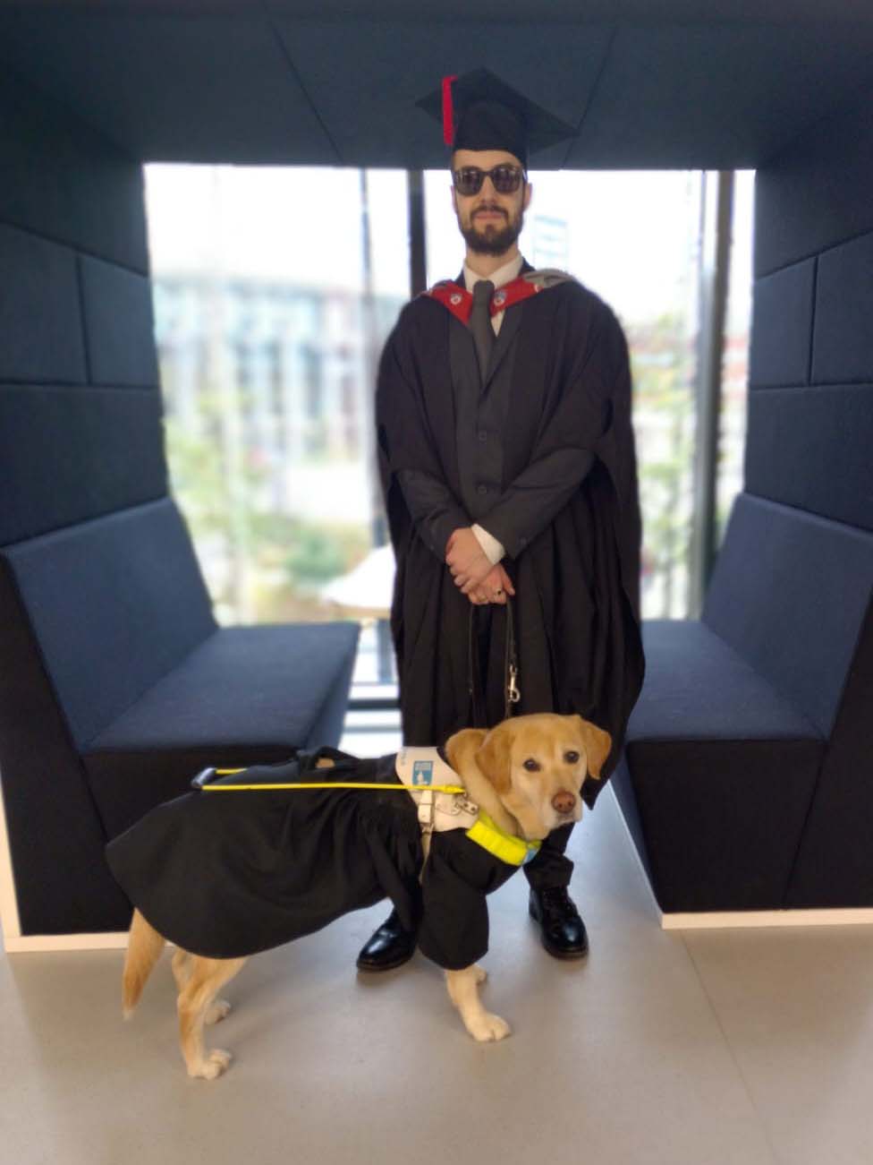 Juan wearing a black mortar board and gown with guide dog Angel standing in front of him wearing a gown